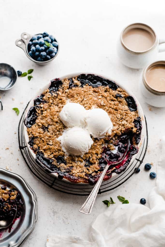 A pie dish of blueberry crisp, topped with vanilla ice cream, with a spoon digging in.