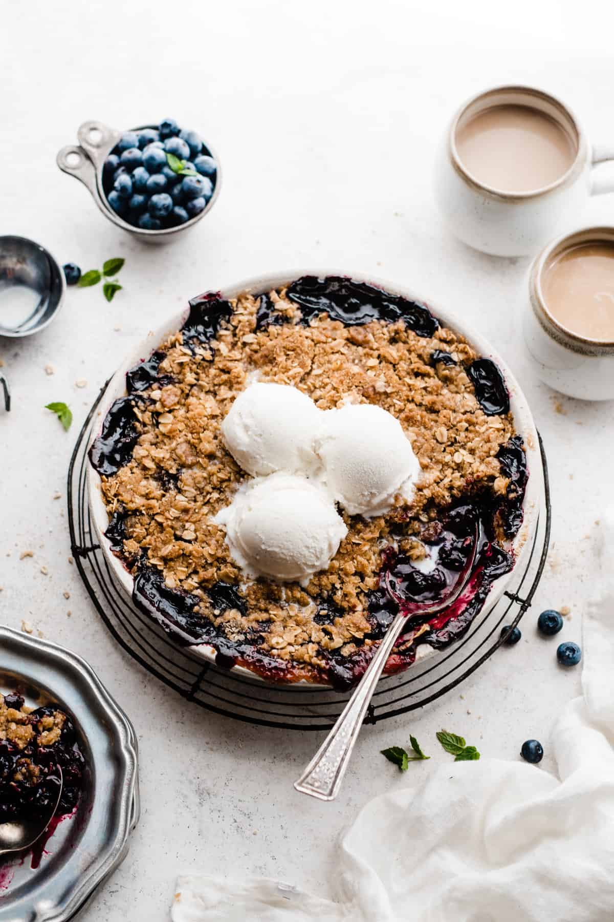 A round dish of blueberry crisp topped with ice cream, with a serving spoon digging in.