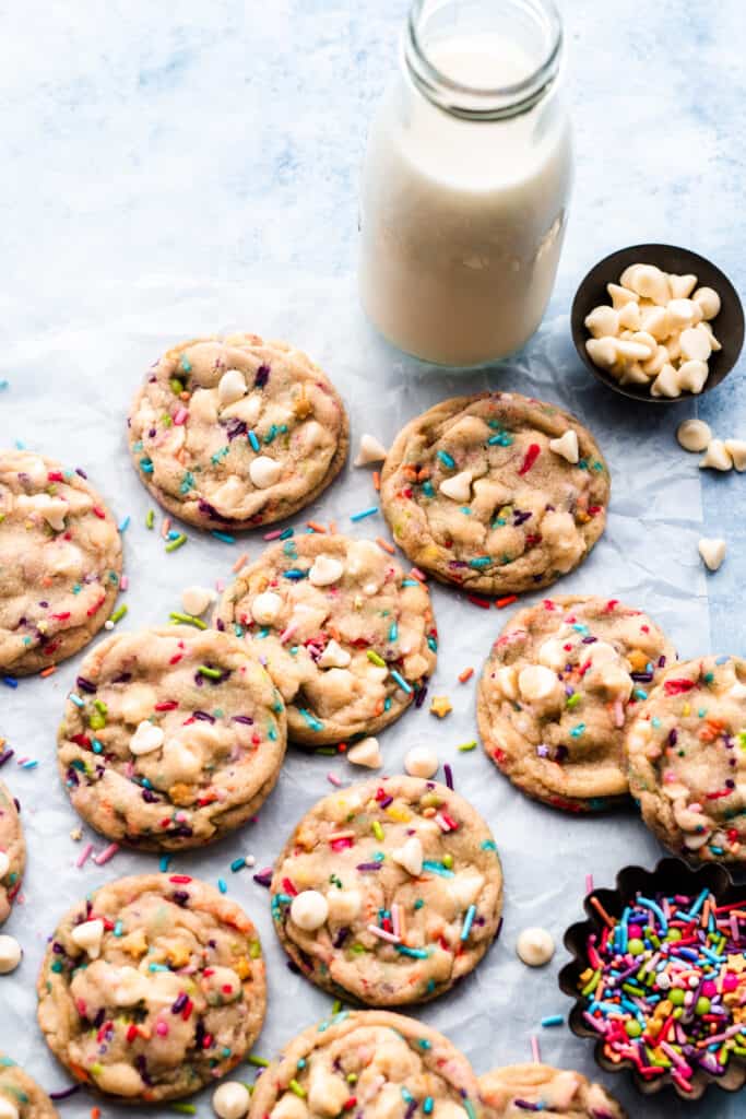 Colorful funfetti cookies on parchment paper with a bottle of milk and small bowls of sprinkles and white chocolate chips.