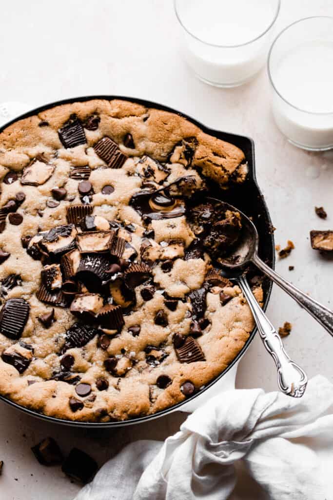 The skillet cookie topped with peanut butter cups and chocolate chips, with spoons scooping bites out.