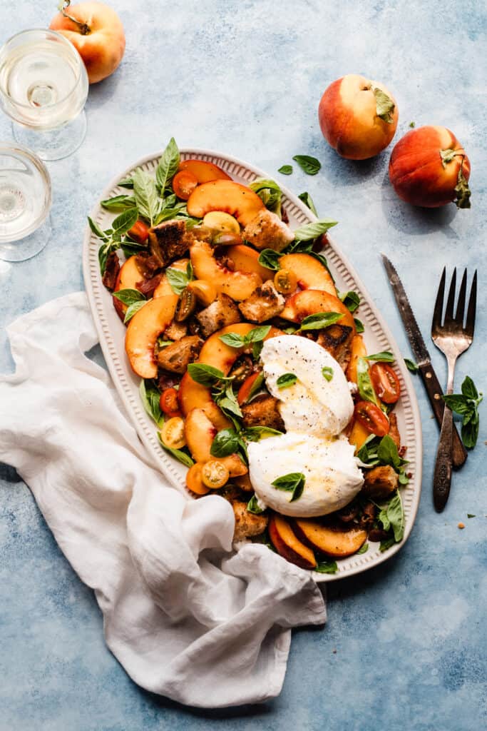 A platter of peach panzanella salad on a blue surface, surrounded by glasses of wine and fresh peaches. 