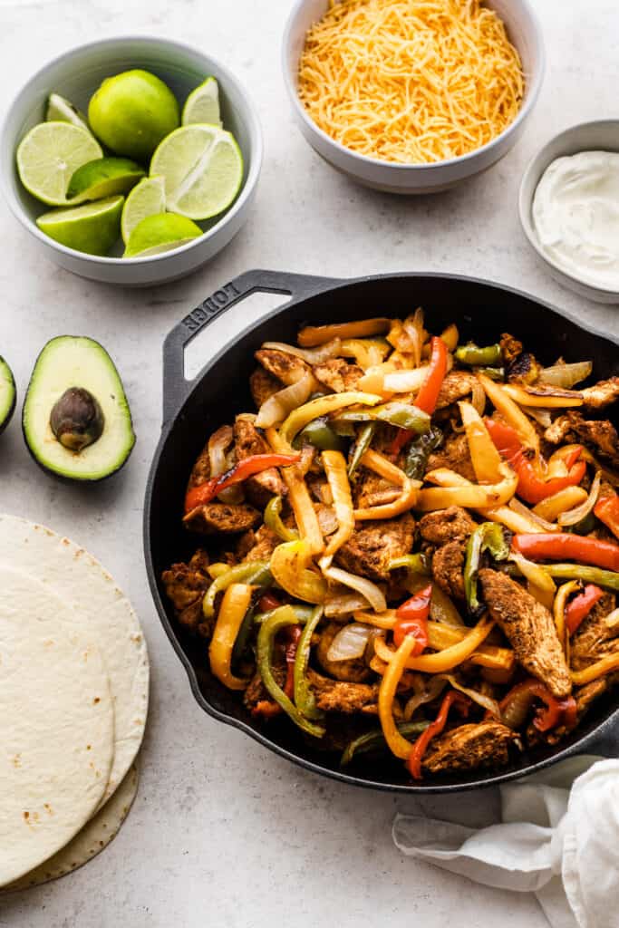 A cast iron skillet full of chicken fajitas, surrounded by bowls of limes, cheese, sour cream, tortillas, and an avocado.