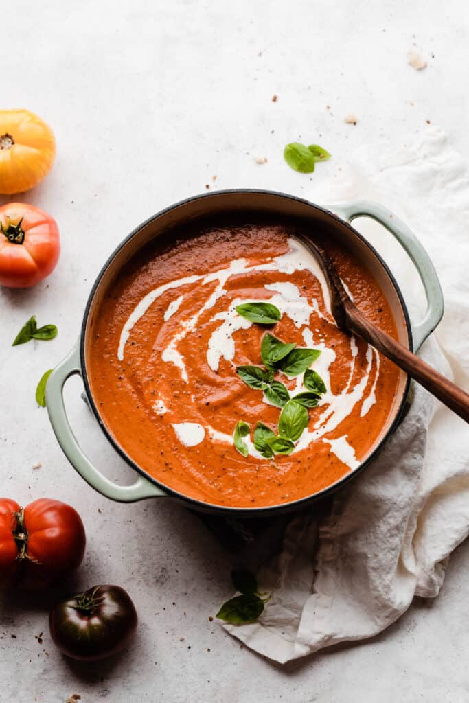A dutch oven filled with bright tomato soup with a drizzle of cream and a handful of fresh basil.