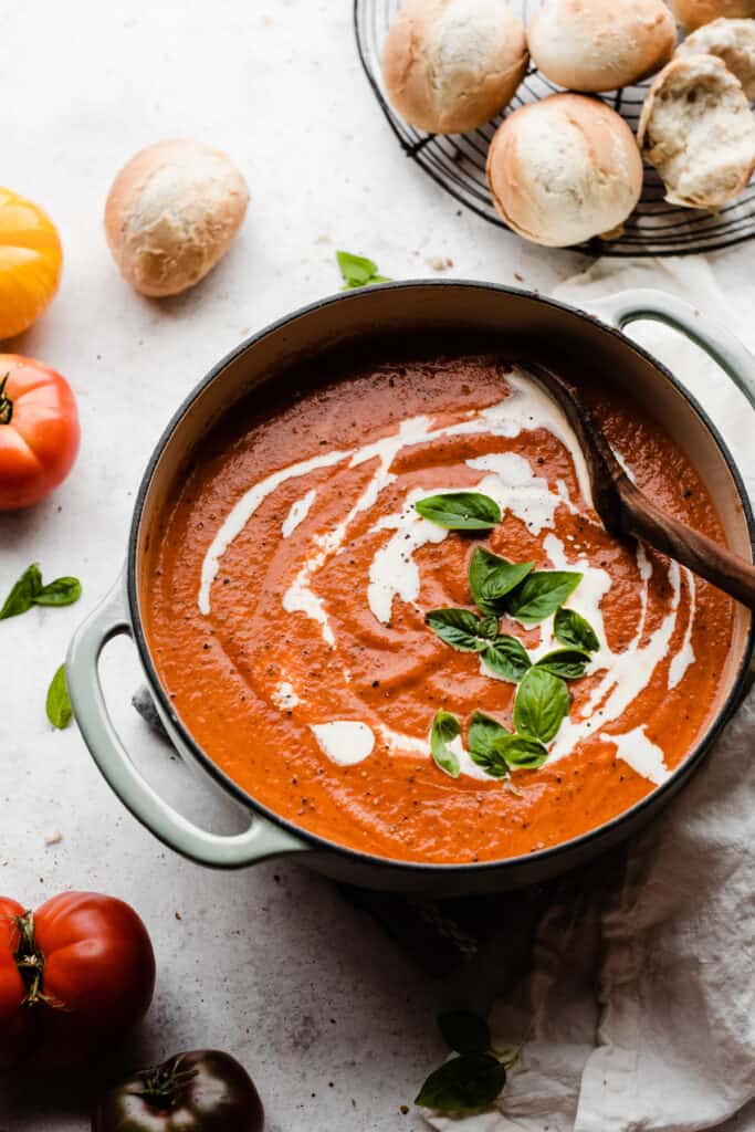 A pot of vibrant tomato basil soup with a tray of rolls nearby.