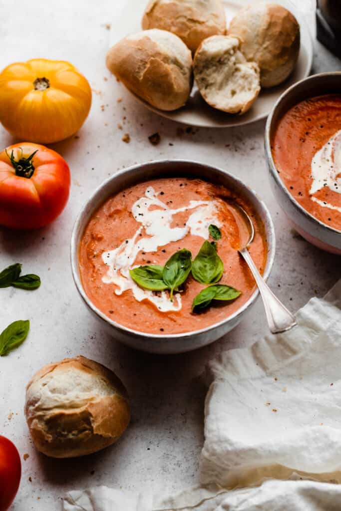 Two bowls of tomato soup drizzled with cream and topped with fresh basil, with a plate of rolls nearby.