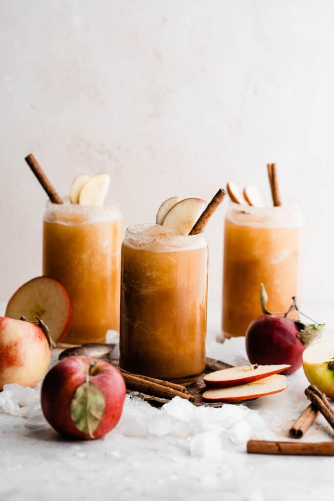 Three glasses of apple cider slushies garnished with apple slices and cinnamon sticks, with ice cubes and apples scattered around.
