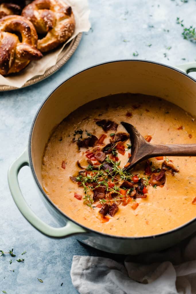 A pot of beer cheese soup topped with bacon, thyme, and red bell pepper.