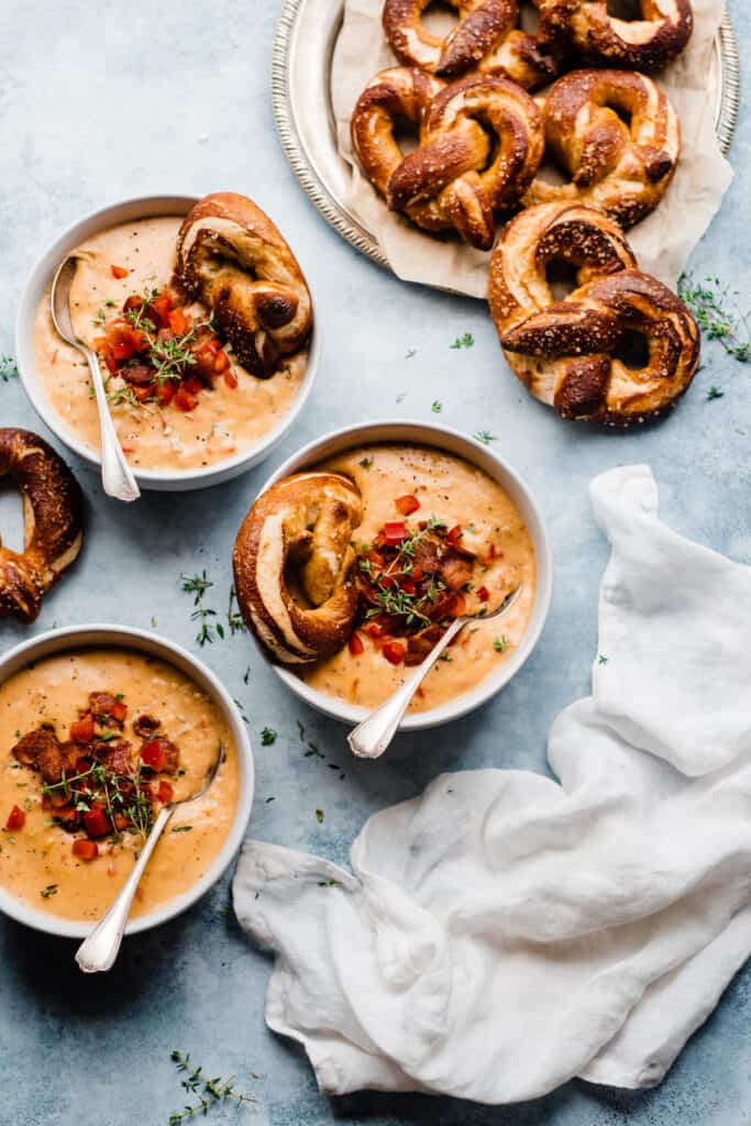 Bird's eye view of bowls of beer cheese soup topped with red pepper, thyme, bacon, and soft pretzels for dunking.