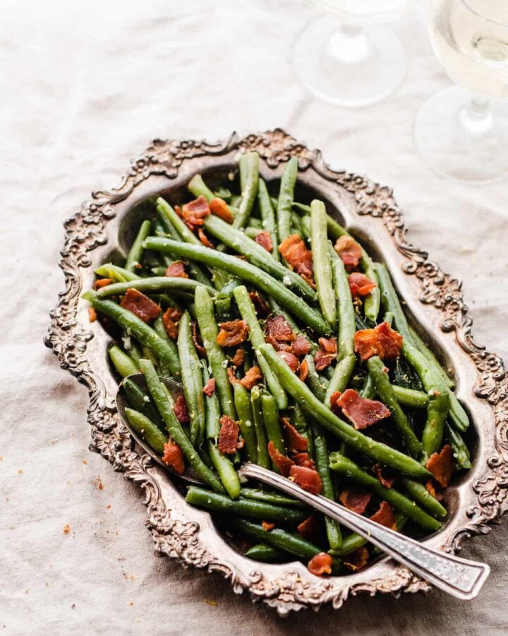 A metal dish filled with bacon green beans on a white linen surface.