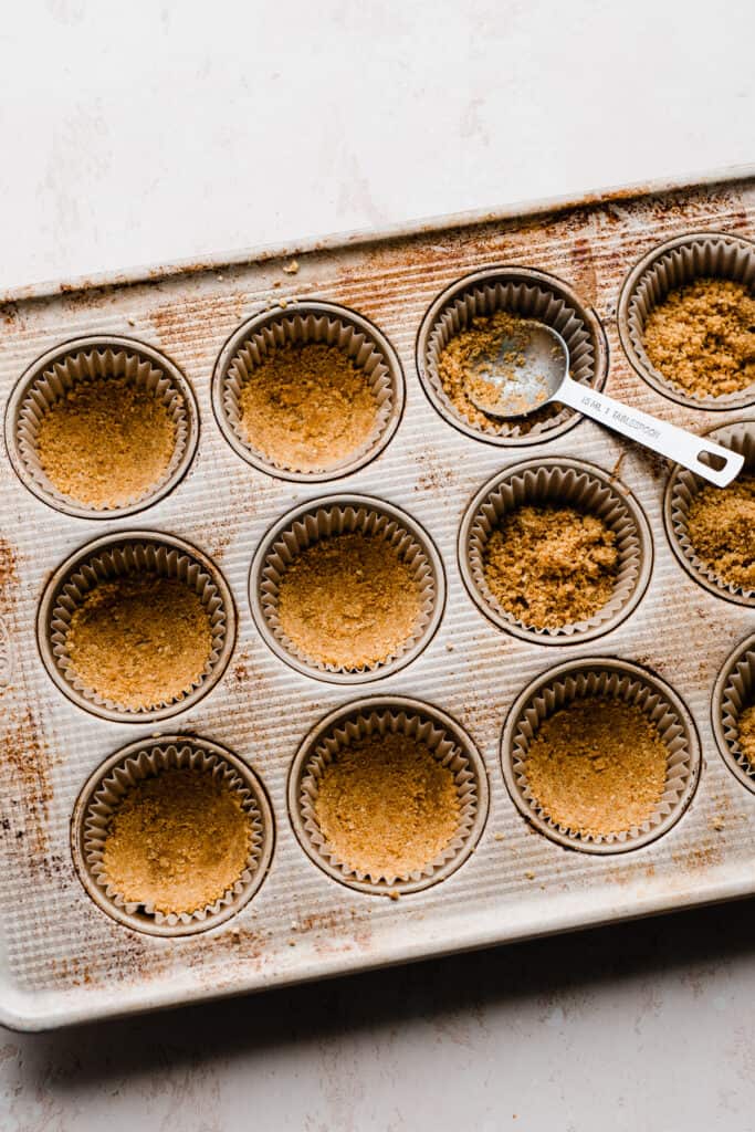 Graham cracker crust being pressed into a muffin pan.
