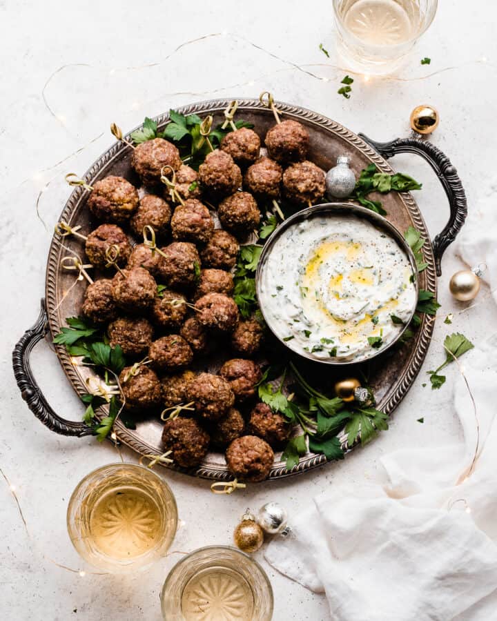 A platter of meatballs on skewers, with a bowl of yogurt dip and glasses of wine.