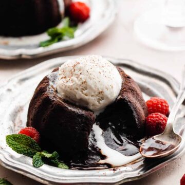 A chocolate lava cake on a plate, topped with ice cream and raspberries, with the molten center spilling out.