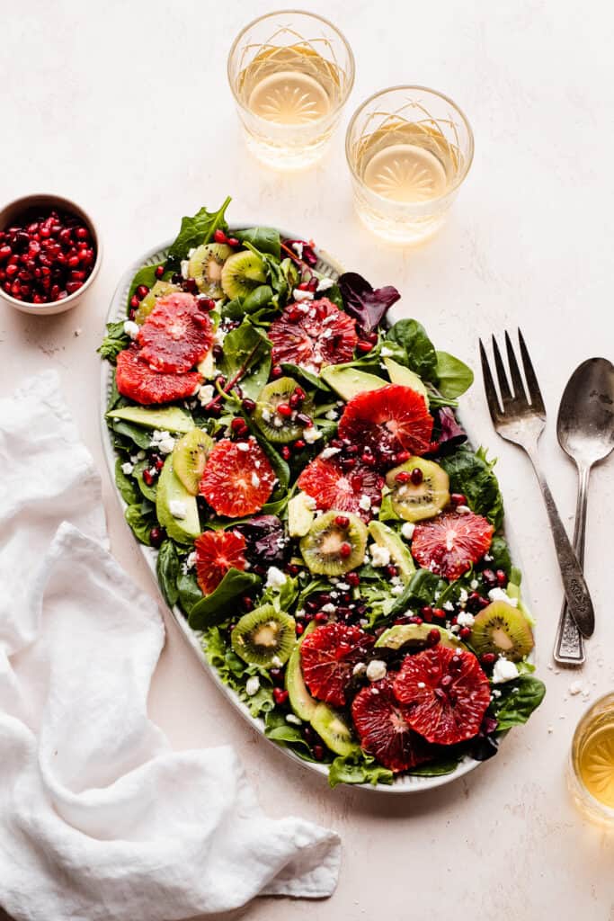 A platter of the colorful salad on a light pink surface, with glasses of wine.