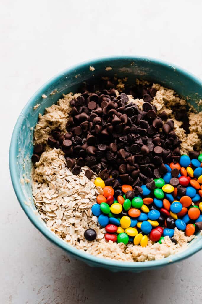 A mixing bowl filled with cookie dough, with m&ms, chocolate chips, and oats on top.