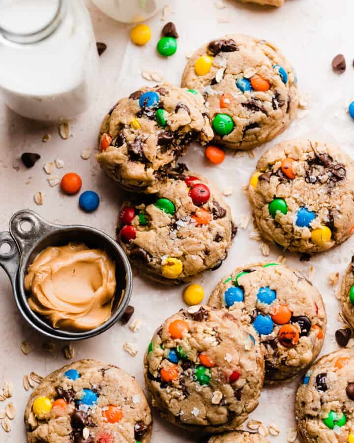 Monster cookies on a light surface, with a bowl of peanut butter and glasses of milk.