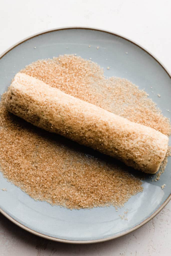 A log of dough being rolled in raw sugar on a plate.