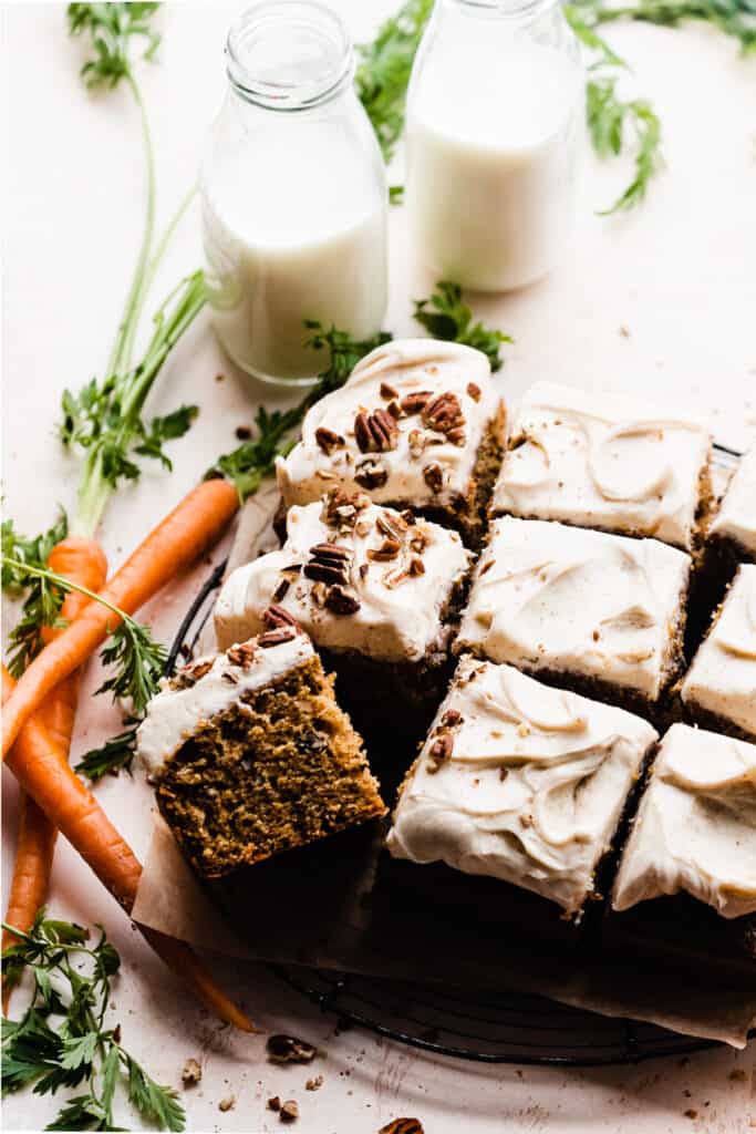 A sliced carrot sheet cake topped with cream cheese frosting and chopped pecans, with two bottles of milk.