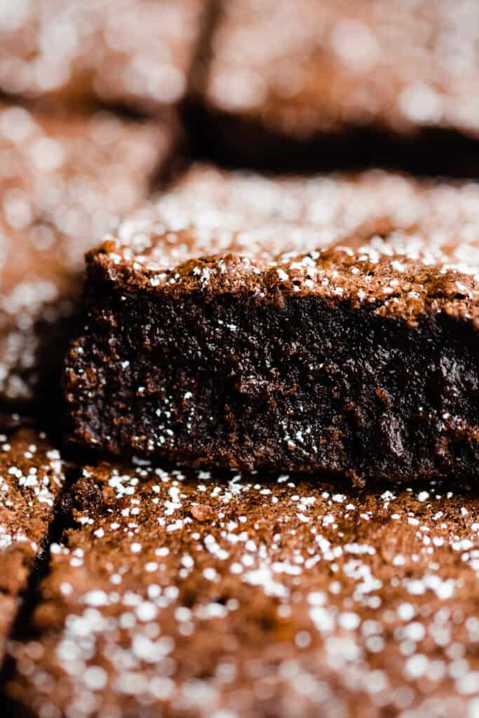 A close-up on a fudgy brownie peeking up out of a row of them, with a dusting of powdered sugar on top.