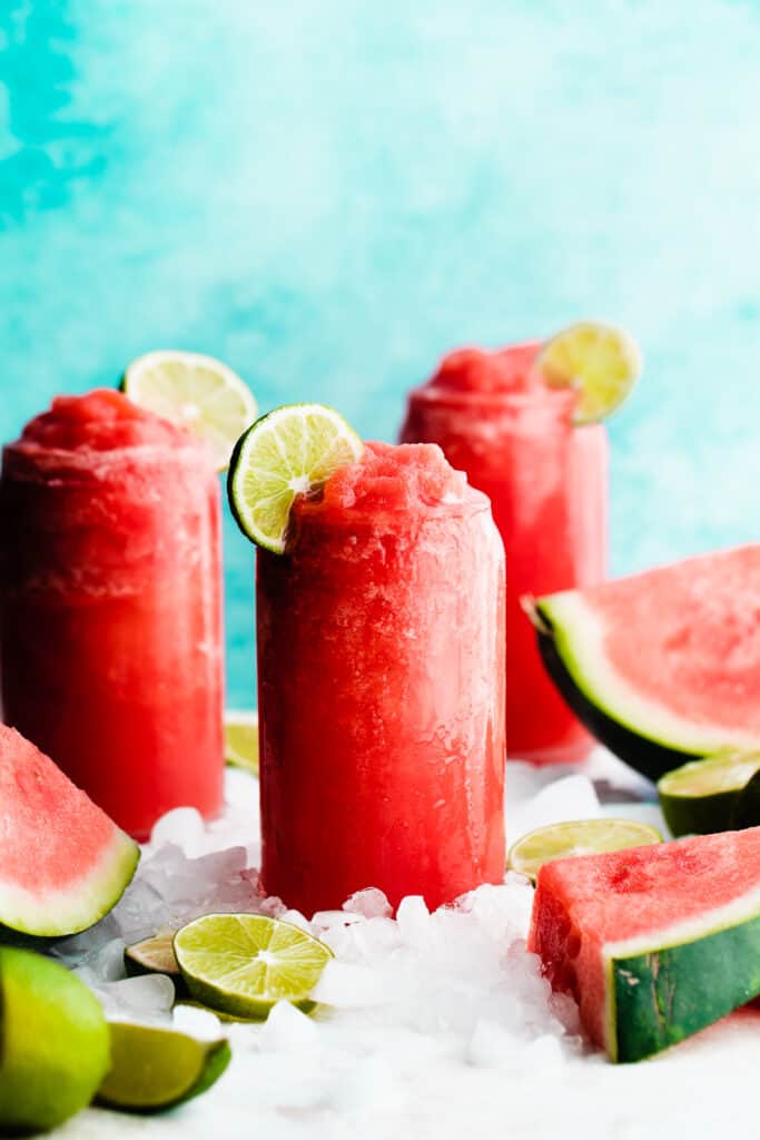 Three glasses of frozen watermelon margaritas surrounded by ice and topped with lime slices.