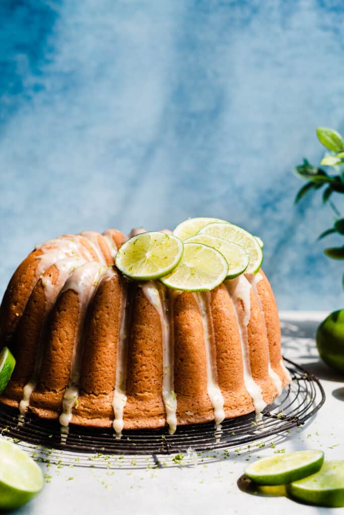 Key Lime Bundt Cake with lime glaze and lime slices on top, on a round rack with a blue backdrop.