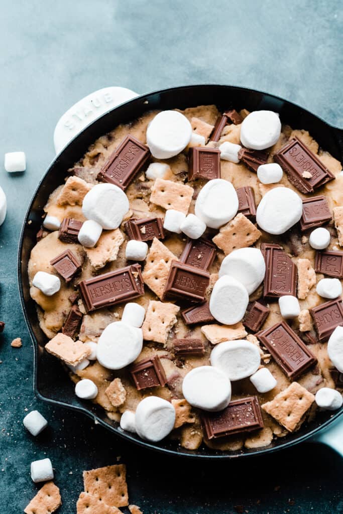 The unbaked s'mores skillet cookie topped with marshmallows, graham crackers, and chocolate pieces.