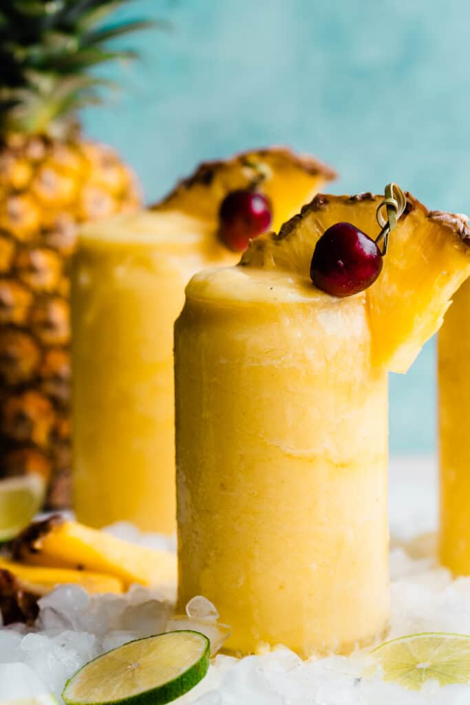 A close-up on a glass of bright yellow pineapple piña colada with a wedge of pineapple and cherry on top.