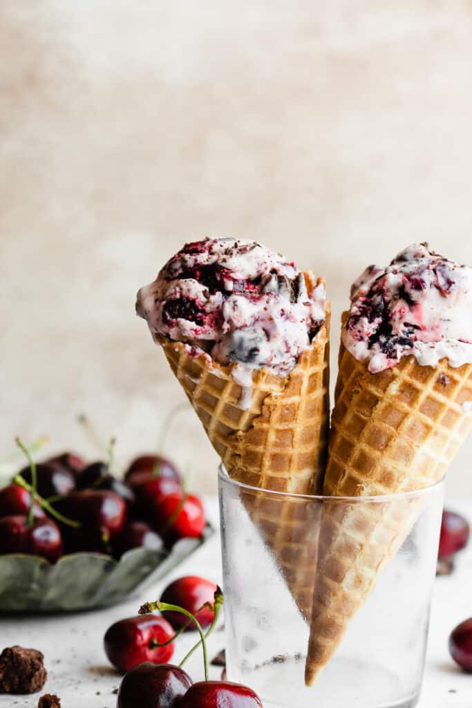 Two cones with black forest ice cream in a glass, with fresh cherries scattered around.