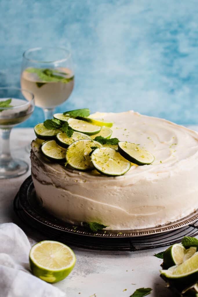 The Mojito Cake on a cake plate with fresh limes and mint on top, against a blue backdrop.