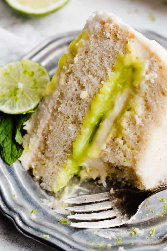 A close up of a slice of lime cake on a plate with light fluffy texture, whipped frosting, and lime curd filling.
