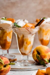 Three peach shakes in old fashioned glasses with peach sauce swirled in and peaches and whipped cream on top.