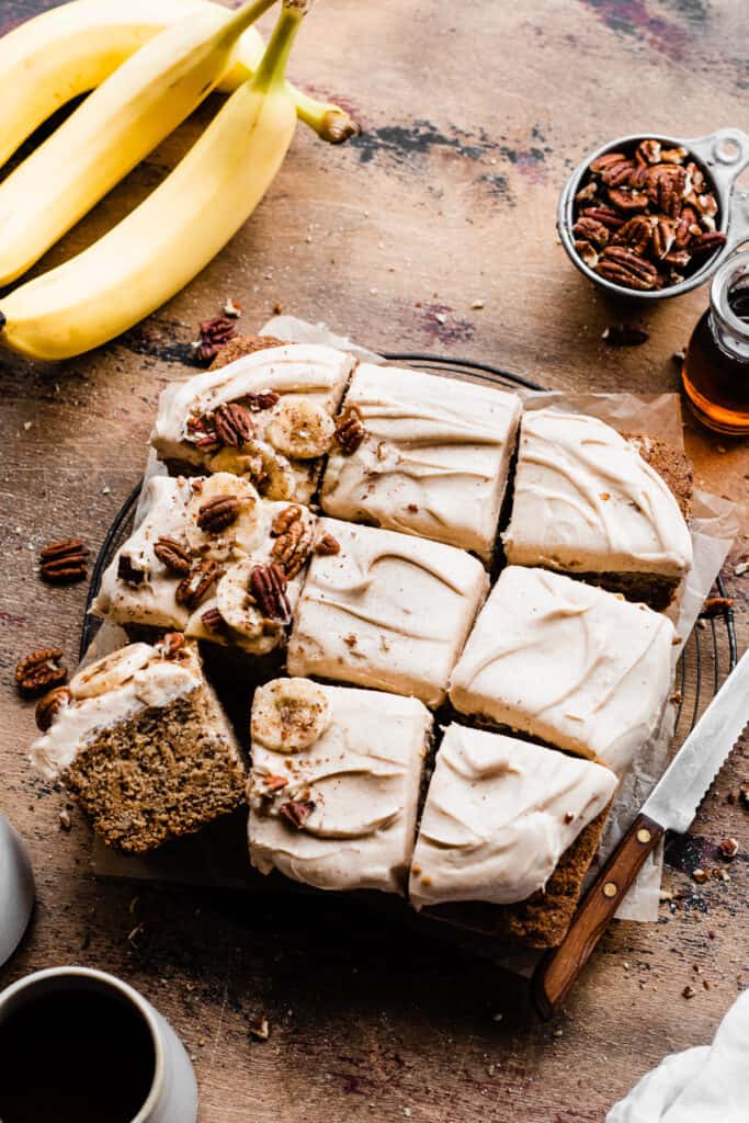 Sliced banana cake on a wooden surface, with bananas, pecans, and a bottle of maple syrup.