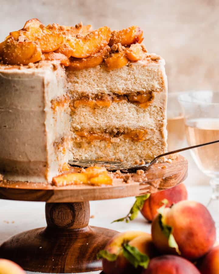 A sliced peach cobbler cake on a cake stand with visible layers of brown butter frosting, peach filling, and streusel.