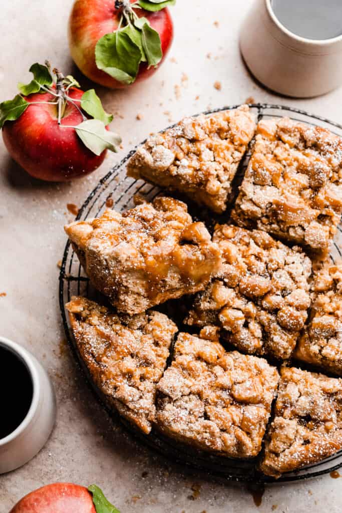 The sliced caramel apple crumb cake on a wire rack, with leafy apples and mugs of coffee nearby.