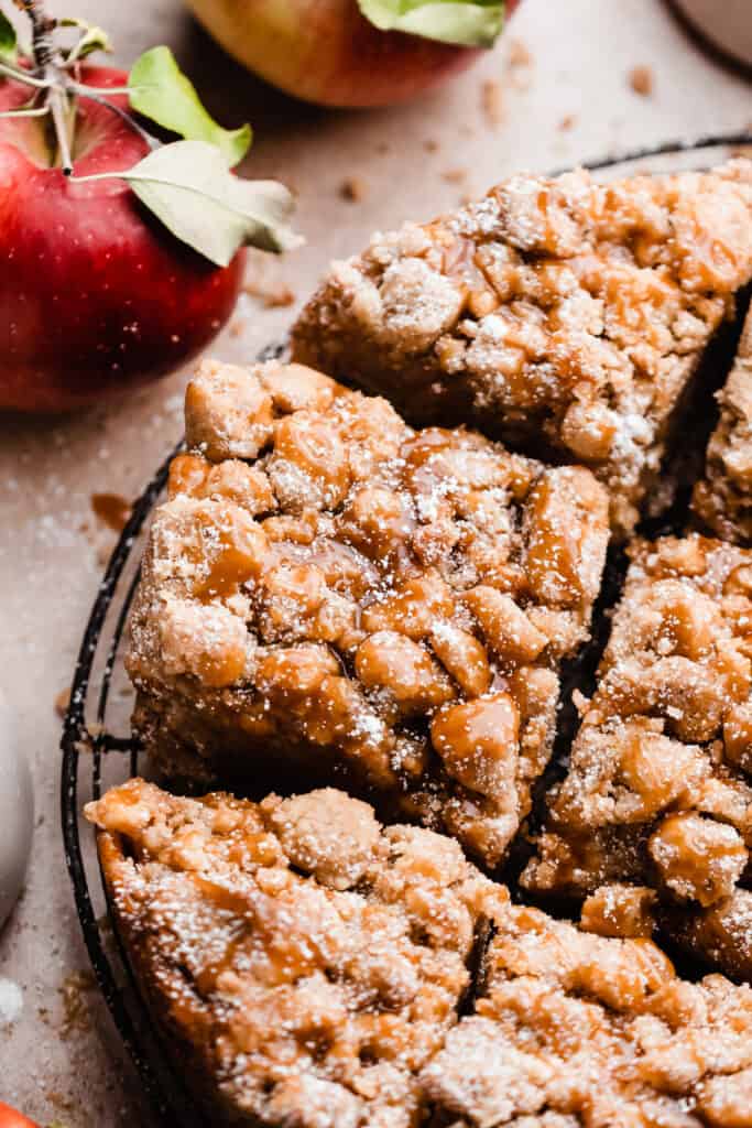 A close-up on slices of the caramel apple crumb cake on a wire rack, with leafy apples nearby.