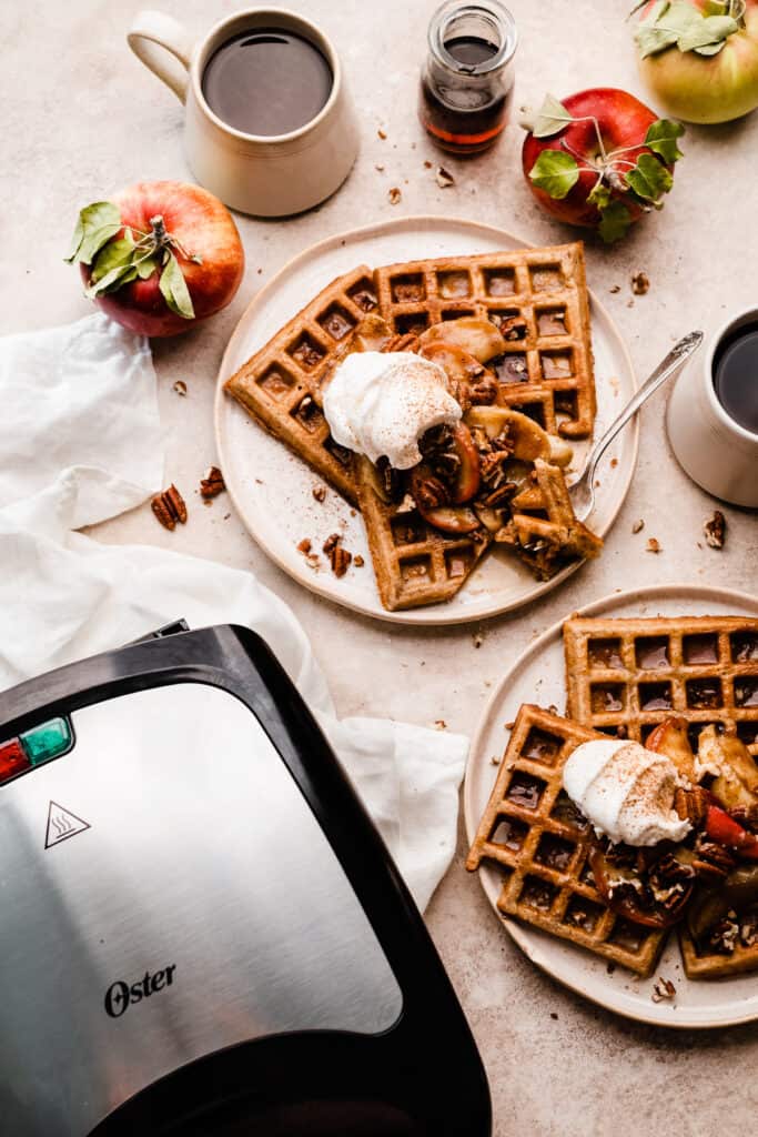 Plates of cinnamon caramelized apple waffles with a waffle maker nearby.