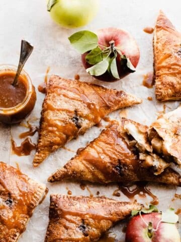 Puff pastry apple turnovers on a light surface with salted caramel sauce drizzled on and whole apples around.