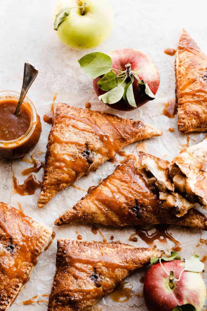 Puff pastry apple turnovers drizzled with salted caramel sauce on a brown surface with whole apples sitting around.