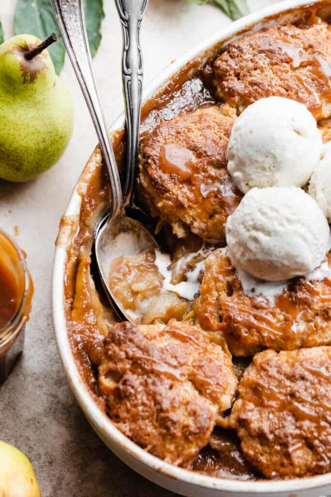 A close up of a spoon digging into the pear cobbler, with melty streams of vanilla ice cream running into the filling.