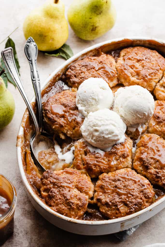 A baking dish of pear cobbler with ice cream on top and a serving spoon digging in.