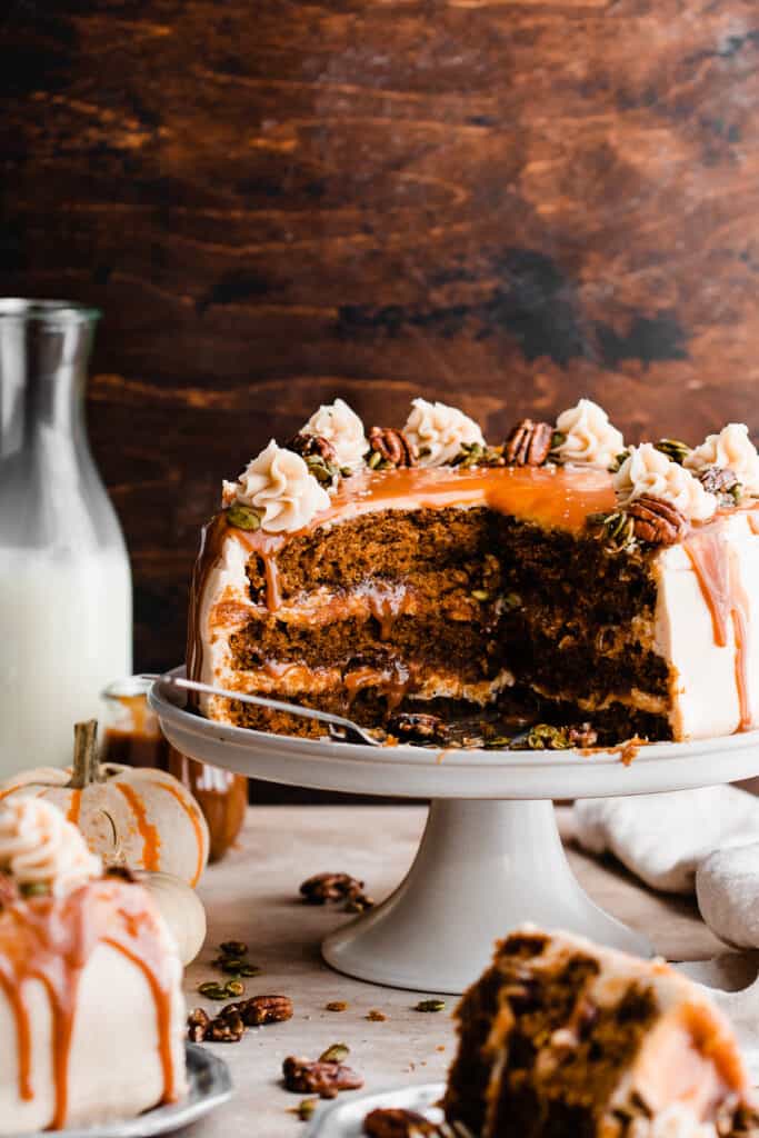 The sliced open cake on a cake stand, showing the layers of fluffy pumpkin cake, cream cheese frosting, candied nuts, and caramel sauce.