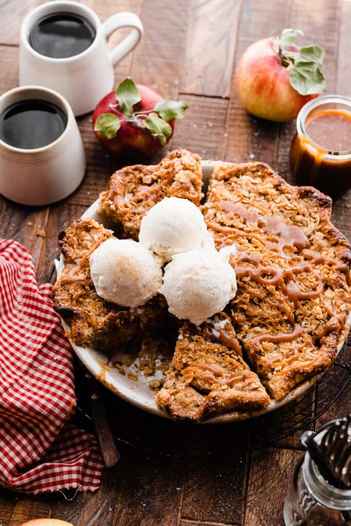 The sliced caramel apple pie topped with salted caramel sauce and vanilla ice cream on a wooden surface, with mugs of coffee.