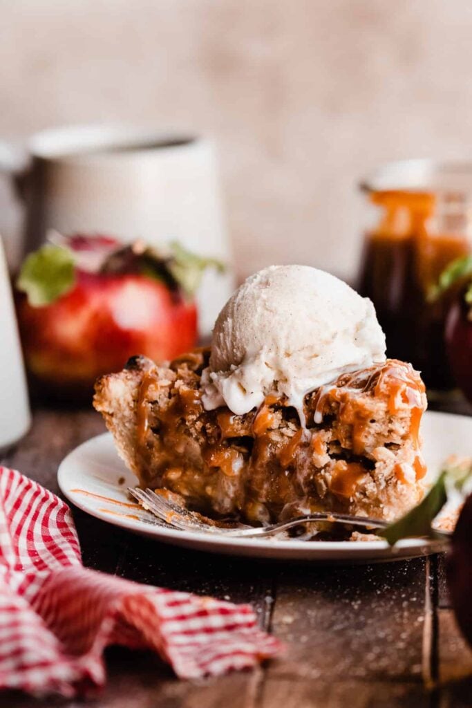 A slice of apple pie baked inside buttery, flaky homemade pie crust, with caramel sauce and a scoop of ice cream.
