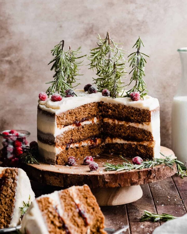 Sliced gingerbread cake on a wooden stand, with sugared cranberries and rosemary trees on top.
