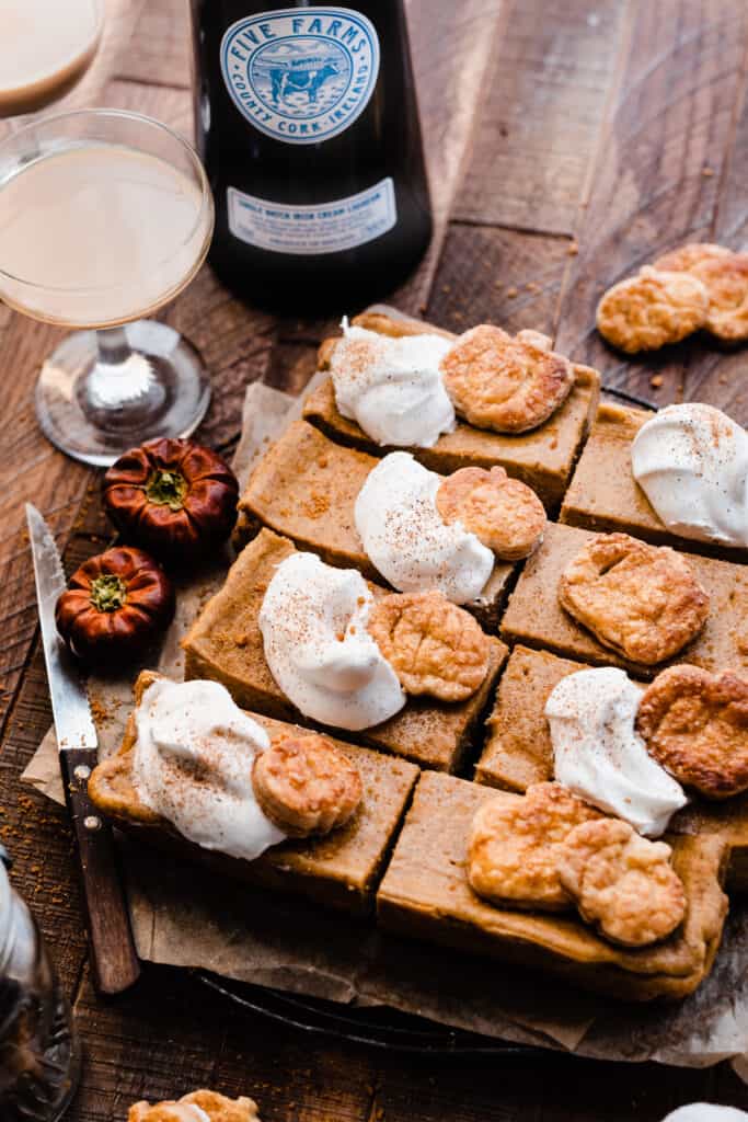 A birds eye view of the sliced pumpkin bars topped with whipped cream on a wooden surface, with glasses of irish cream.