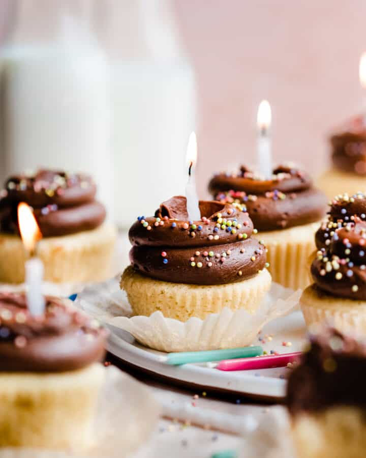 Yellow birthday cupcakes with chocolate frosting and rainbow sprinkles with lit birthday candles.