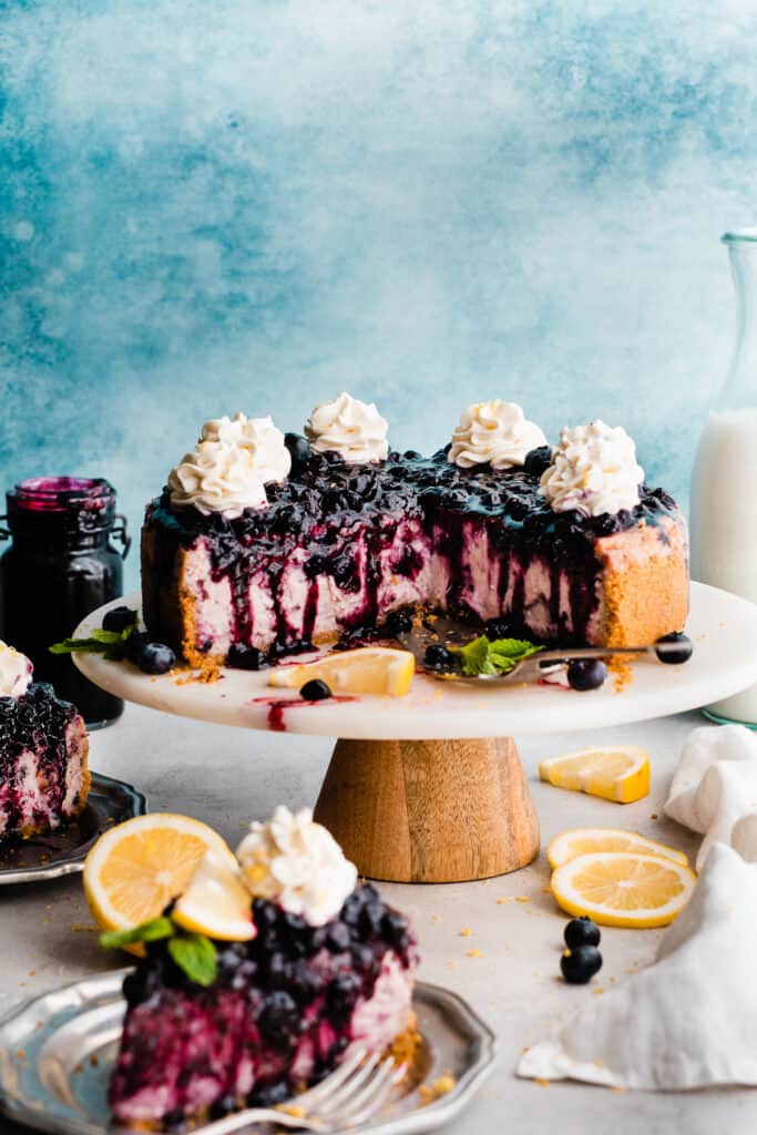 The sliced cheesecake on a cake stand with whipped cream swirls and blueberry sauce on top.