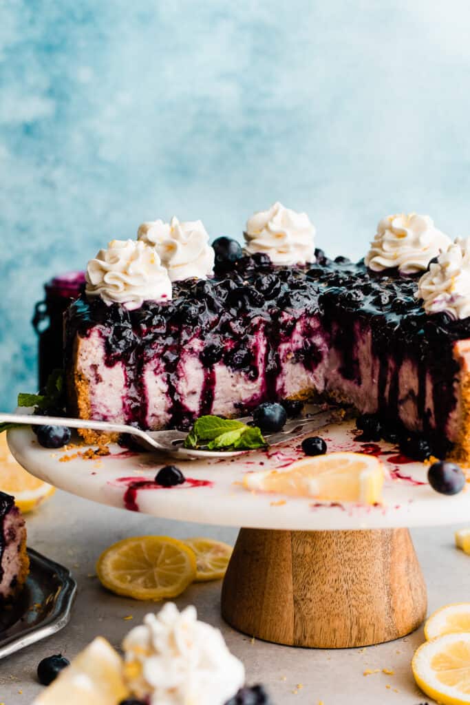 The sliced cheesecake on a cake stand with whipped cream swirls and blueberry sauce on top.
