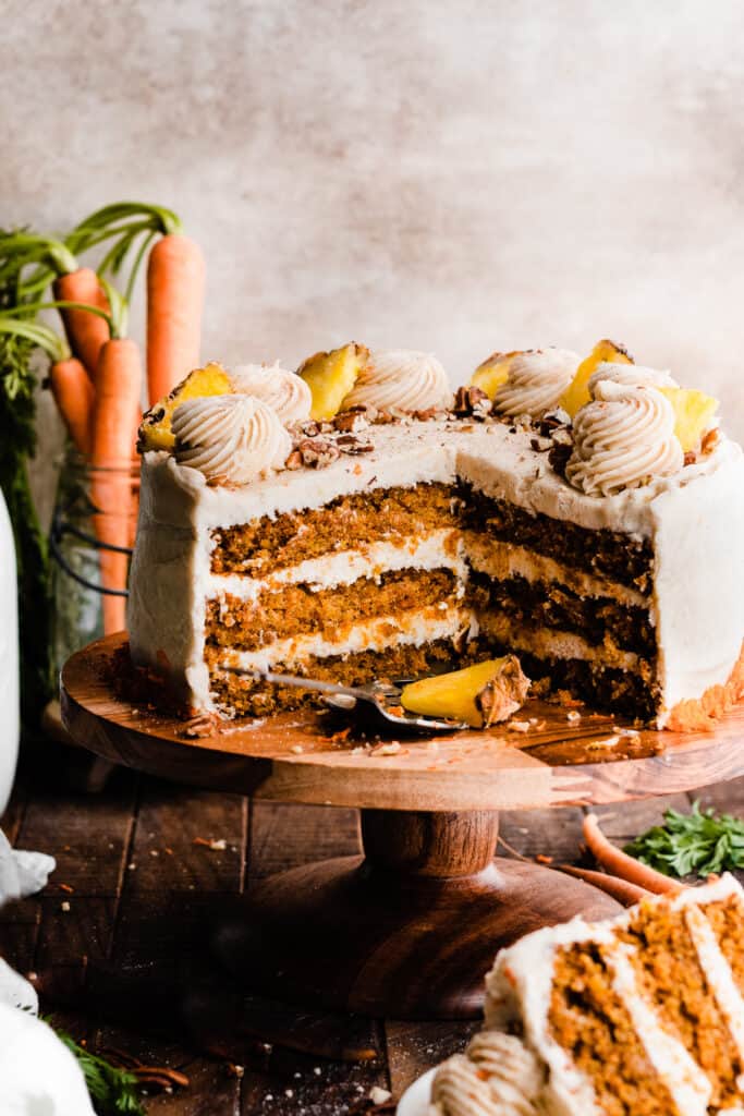 The sliced open cake on a cake stand, with the visible layers of fluffy cake and carrots in the background.