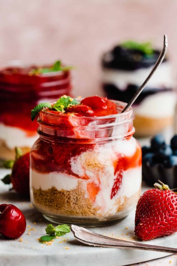 Jars of mini cheesecakes, with a spoon digging into the strawberry topped cheesecake.