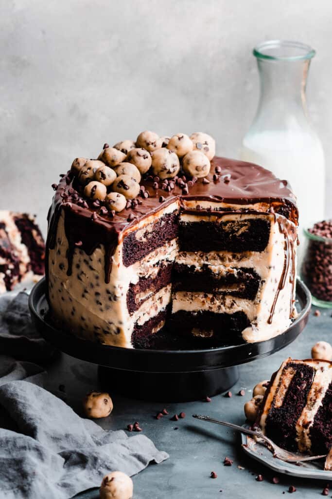 The sliced open cookie dough cake on a cake stand, topped with ganache and cookie dough balls, with a bottle of milk in back.
